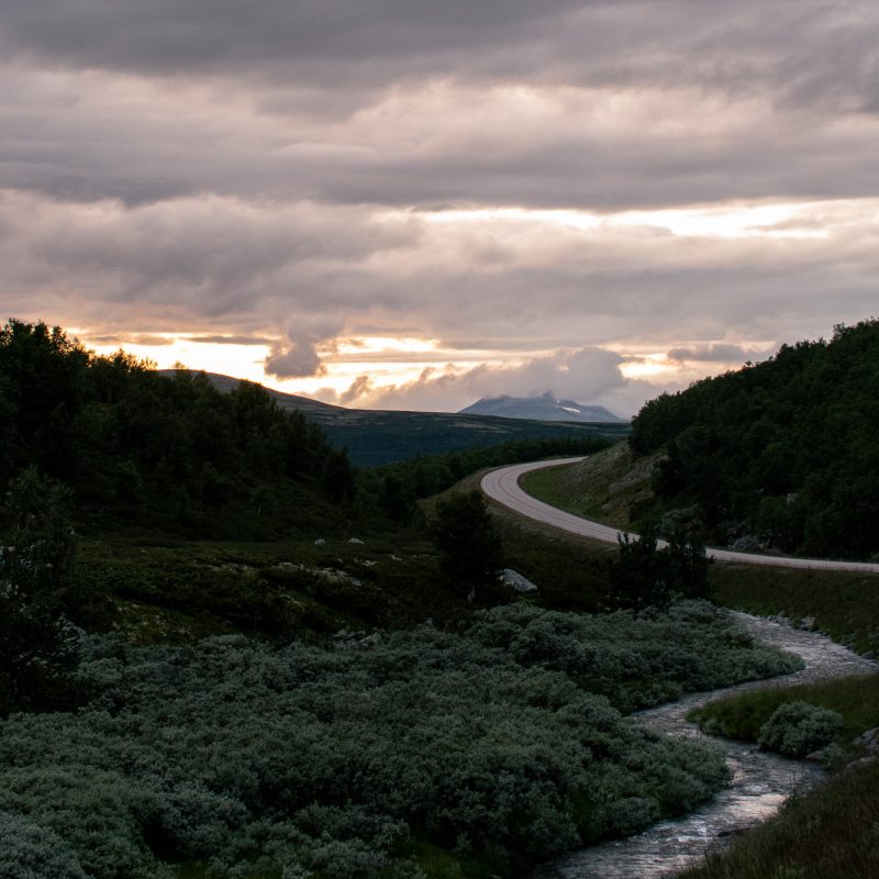 **Titel** *Tegen het Licht van de Avond* **Omschrijving** Dit beeld is gemaakt op het moment dat de dag langzaam kantelt richting avond. De zon staat laag, grotendeels verborgen achter wolken en bergen, maar laat nog net genoeg licht door om het landschap richting te geven. De weg slingert het dal in en fungeert als stille gids door het beeld, terwijl het bergstroompje op de voorgrond koel en ingetogen blijft, als tegenwicht voor de warme gloed aan de horizon. De kracht van deze foto zit niet in spektakel, maar in balans. Het contrast tussen warm en koel, tussen licht en schaduw, en tussen beweging en stilte zorgt voor een gelaagd beeld dat rust uitstraalt zonder statisch te worden. De lucht draagt het verhaal, het landschap ondersteunt, en de voorgrond verankert de kijker in het moment. Dit werk nodigt uit tot langzaam kijken. Het past goed in interieurs waar ruimte is voor nuance, natuurlijke tinten en een kalme, contemplatieve sfeer.