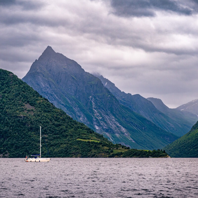 Zeilboot op een Noors fjord, omringd door steile groene bergen onder een bewolkte lucht.